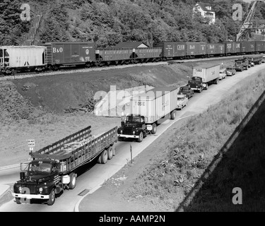 1950 LIGNE DE TRAFIC AVEC DE NOMBREUX CAMIONS ET VOITURES & FREIGHT train passant sur une colline adjacente Banque D'Images