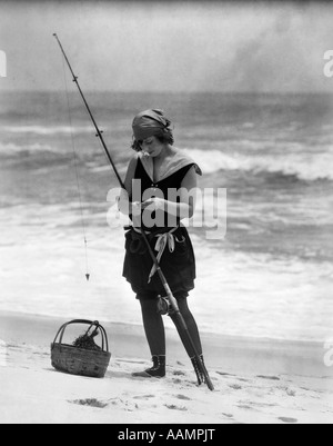 Années 1920 FEMME EN MAILLOT COSTUME STANDING ON BEACH PUTTING APPÂT SUR POTEAU DE PÊCHE SURF Banque D'Images