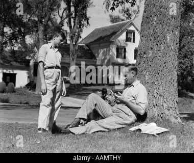 Années 1930 Années 1940 PÈRE ET CHIEN assis sous arbre fils adolescent se tenant debout dans une cour Banque D'Images