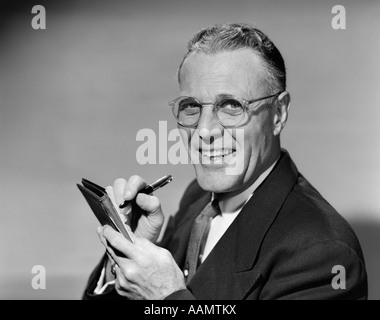 1950 HOMME MÛR DANS LE PORT DE LUNETTES RÉPONDRE À TENIR PEN ÉCRIT DANS L'ORDINATEUR PORTABLE LOOKING AT CAMERA Banque D'Images