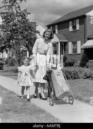 1950 MÈRE LOOKING AT CAMERA SMILING MARCHANT SUR UN TROTTOIR DE BANLIEUE HOLDING DAUGHTER'S HAND HELPING SON PANIER D'ÉPICERIE POUSSANT Banque D'Images