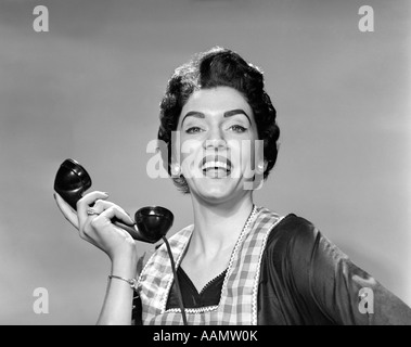 1950 WOMAN IN APRON HOLDING RÉCEPTEUR DU TÉLÉPHONE EN MAIN, LOIN DE l'oreille à la caméra en souriant Banque D'Images