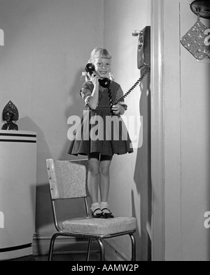 Années 1960 Années 1950 JEUNE FILLE DEBOUT SUR CHAISE DE CUISINE EN CONVERSATION SUR TÉLÉPHONE MURAL LOOKING AT CAMERA Banque D'Images