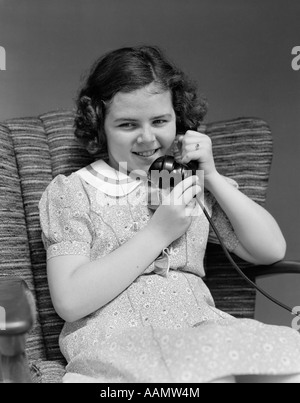 1930 LITTLE GIRL SITTING IN CHAIR TALKING ON TELEPHONE Banque D'Images