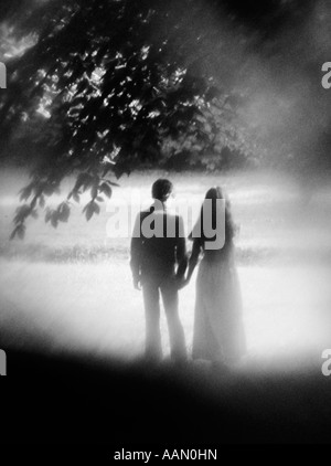 1970 VUE ARRIÈRE SOFT-FOCUS SHOT OF COUPLE STANDING UNDER TREE HOLDING HANDS Banque D'Images