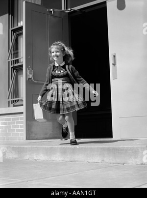 Années 1950 Années 1960 fille blonde COURT PORTE AVANT DU BÂTIMENT DE L'ÉCOLE TENUE LUNCH BOX OU SAC pull robe à carreaux SMILING Banque D'Images