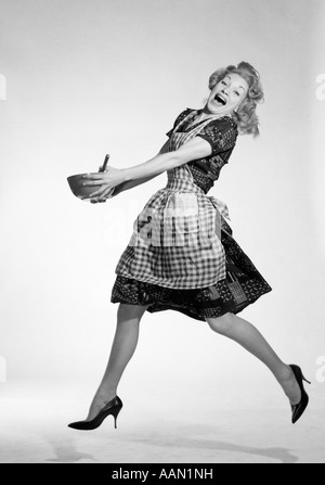 1950 FEMME EN TABLIER À CARREAUX DE SAUTER AVEC ALLÉGRESSE HOLDING SPOON bol à mélanger avec les deux mains Grand sourire Banque D'Images