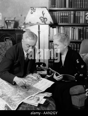 1930 VIEUX COUPLE HOMME FEMME LA LECTURE DE CARTES LIVRES BROCHURES VOYAGE assis par étagère Banque D'Images