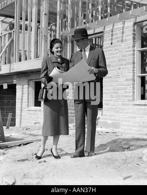 1950 SMILING COUPLE LOOKING AT BUILDING PLANS DEBOUT DEVANT DE MAISON INACHEVÉE Banque D'Images