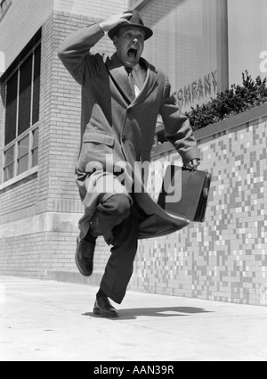 Années 1960 Années 1950 FRANTIC MAN RUNNING DOWN STREET HOLDING HAT SUR LA MAIN AVEC CARRYING BRIEFCASE PORTER TOP COAT Banque D'Images