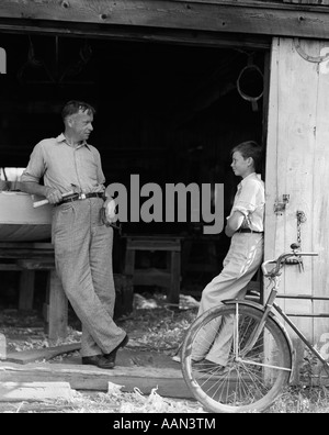 1930 HOMME PÈRE HOLDING HAND TOOLS PARLER À BOY fils se penchant porte de hangar de bateau Banque D'Images