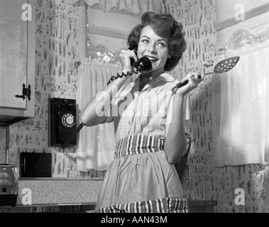 Années 1950 Années 1960 SMILING WOMAN HOLDING SPOON TÉLÉPHONE MURAL Portant tablier, debout dans la cuisine Banque D'Images