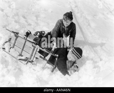 Années 1930 Années 1940 DEUX GARÇONS RIRE DANS LA NEIGE TOMBER DU TRAÎNEAU QUI EST TOURNÉE SUR SON CÔTÉ Banque D'Images