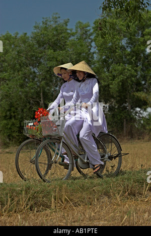 Deux jeunes femmes à chapeaux coniques et ao dai blanc traditionnel costume de la bicyclette sur le chemin des rizières près de Phan Thiet Vietnam Banque D'Images