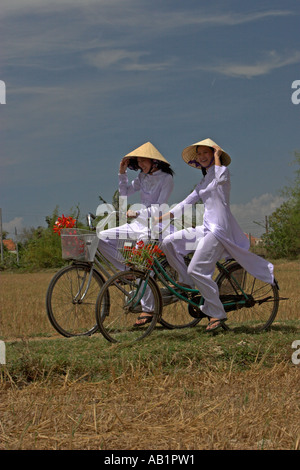 Deux jeunes femmes à chapeaux coniques et ao dai blanc traditionnel costume de la bicyclette sur le chemin des rizières près de Phan Thiet Vietnam Banque D'Images