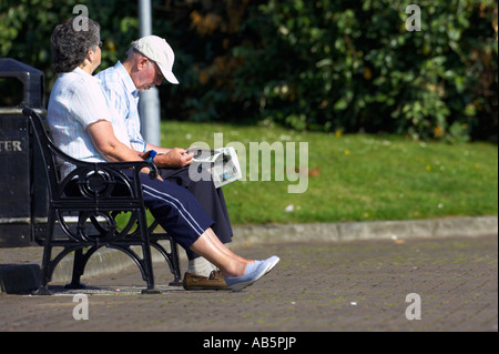 Vieil Homme et femme couple en train de lire un journal sur un banc de parc dans sunshine london Irlande du Nord Banque D'Images