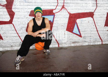 Jeune homme assis sur le basket-ball contre graffiti sur mur de brique Banque D'Images