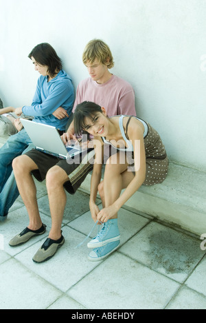 Groupe de jeunes amis assis sur un banc, une jeune femme attachant lacets and smiling at camera Banque D'Images