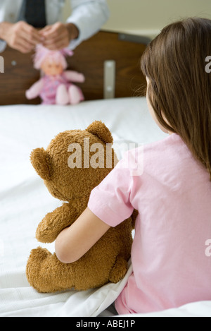Girl sitting in hospital bed holding teddy bear, en face de doctor holding doll Banque D'Images