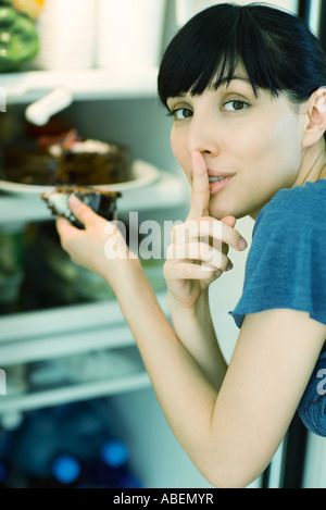 Woman taking part de gâteau du réfrigérateur, looking at camera avec le doigt sur les lèvres Banque D'Images