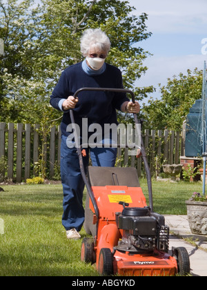 Femme senior fauchant la pelouse domestique du jardin avec une tondeuse à gazon tout en portant un masque anti-poussière pour éviter la fièvre du foin. Royaume-Uni Banque D'Images