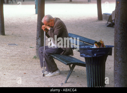 Homme âgé assis seul sur un banc de parc à Paris, la tête baissée appuyée sur son bâton de marche. Vieilli, pauvre, adulte solitaire. France Banque D'Images