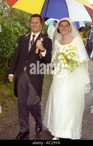 Bride & Groom sortant de l'église portant un parapluie sous la pluie Banque D'Images