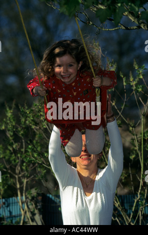 Entraînée par sa mère, une petite fille d'avoir un rendez-vous sur une balançoire. Filette jouant sur une balançoire aidée par sa maman. Banque D'Images