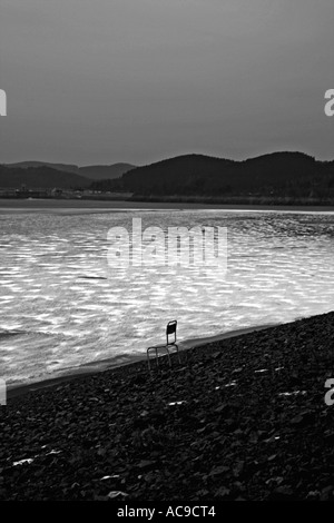 Une chaise solitaire face à un lac calme, entourée de rivages rocheux et de collines lointaines, en noir et blanc. Banque D'Images