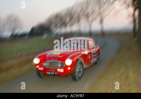 Trois quart avant de voir l'Austin Healey 100/6 Rouge voiture rallye en vitesse sur route goudronnée bordée d'au crépuscule avec des stries de mouvement Banque D'Images