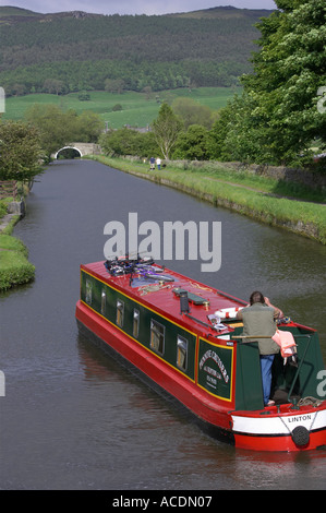 Red & green 15-04 (voitures) bateau naviguant sur le canal de Leeds Liverpool rural pittoresque sur la journée ensoleillée, 1 homme debout par timon - North Yorkshire, England, UK Banque D'Images