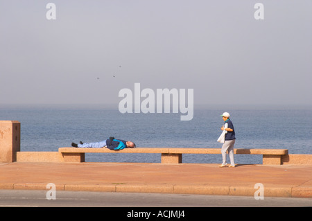 Une femme marche sur le trottoir avec une thermos d'eau chaude ballon avec mate tisane et un homme couché sur un banc, dormir sur la promenade de bord de rivière le long du fleuve Rio de la Plata sur Las Ramblas, Gran Bretagna et Republica Argentina Montevideo, Uruguay, Amérique du Sud Banque D'Images