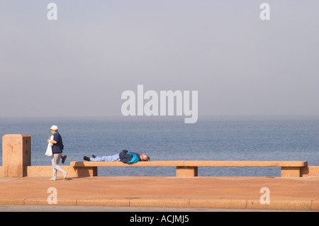 Une femme marche sur le trottoir avec une thermos d'eau chaude ballon avec mate tisane et un homme couché sur un banc, dormir sur la promenade de bord de rivière le long du fleuve Rio de la Plata sur Las Ramblas, Gran Bretagna et Republica Argentina Montevideo, Uruguay, Amérique du Sud Banque D'Images