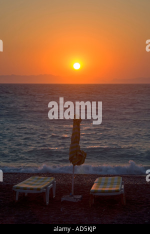 Vue verticale d'une belle couleur orange coucher du soleil sur la mer Egée avec chaises longues et parasols vide sur la plage. Banque D'Images