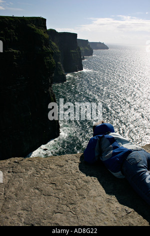 Une jeune femme sur son ventre prend dans la vue de les Falaises de Moher du rocher plat l'affichage au-delà de la barrière Banque D'Images