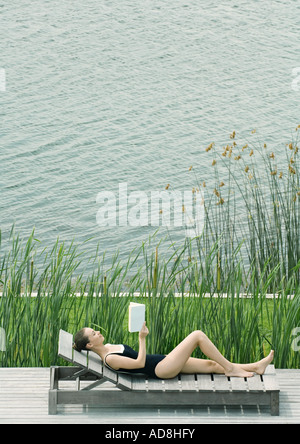 Femme couchée sur une chaise longue, d'une lecture, à côté du lac Banque D'Images