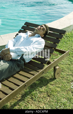 Businessman position allongée sur une chaise longue près de la piscine, holding laptop on lap Banque D'Images