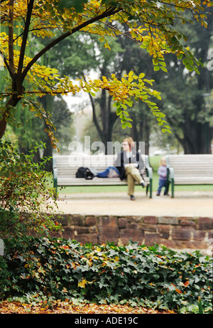 Vue éloignée à travers feuilles et mur de vieille femme assis sur un banc en automne avec tout-petit se tiennent à proximité des jardins du Trésor Banque D'Images