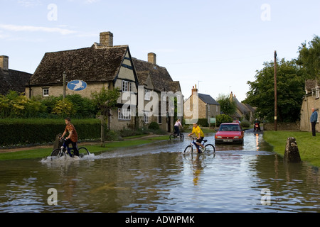 Les enfants à prendre leur vélo à travers l'eau d'inondation dans les Cotswolds Minster Lovell Oxfordshire England Royaume-Uni Banque D'Images