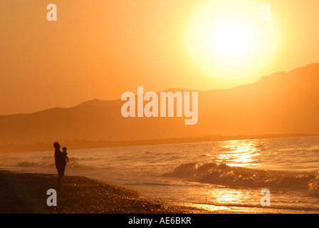 Crète, Grèce. Un parent en tenant son bébé et regarder le coucher de soleil sur la plage entre Maleme et Gerani près de Hania. Banque D'Images