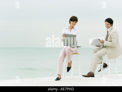 Sur des tabourets Businesspeople sitting on beach, l'un à l'aide d'un ordinateur portable, un autre journal de lecture Banque D'Images