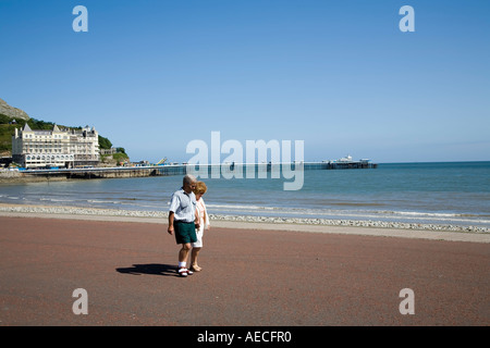 Couple d'âge moyen marche sur la promenade avec le grand hôtel et jetée victorienne dans la distance Llandudno Galles UK Banque D'Images