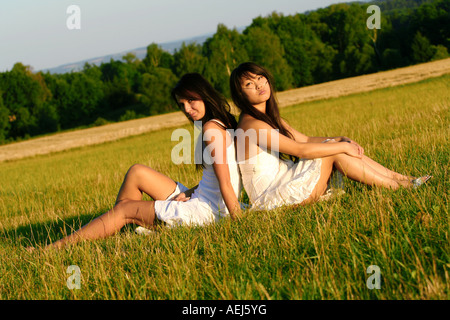 Deux jeunes femmes assises dos à dos sur un terrain herbeux en été, entourées d'arbres et profitant d'une journée paisible et ensoleillée dans la nature. Banque D'Images