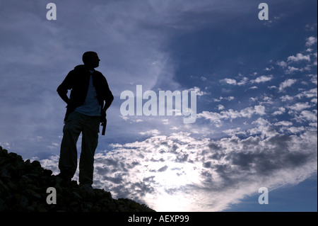 Portrait d'un homme adulte debout sur un tas de roches. Il est découpé sur un fond bleu, ciel nuageux. Plan horizontal. Banque D'Images