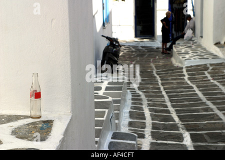 Bouteille verre vide en premier plan avec la moto et un couple assis sur un pas en arrière-plan d'une stone street, isl Mykonos Banque D'Images