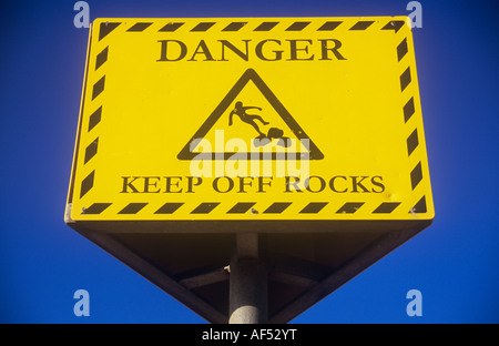 Signe jaune et noir clair contre ciel bleu profond indiquant Danger Garder rocks off avec symbole iconique de l'homme glisser sur les roches Banque D'Images