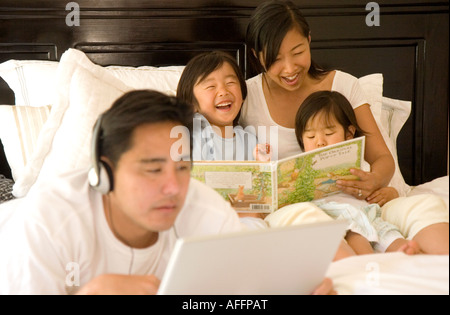 Père écoute au casque branché à l'ordinateur tandis que mère lit aux enfants en arrière-plan Banque D'Images