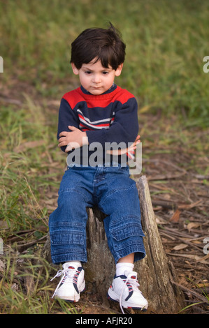 Petit garçon avec les cheveux bruns et un chandail rayé jeans et baskets assis sur une souche d'arbre en colère que si dans le temps à Banque D'Images