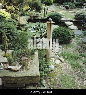 Bonsai arbres dans l'auge de pierre à côté de la fontaine de bambou et lily piscine dans le jardin de style oriental avec des tremplins à travers stream Banque D'Images
