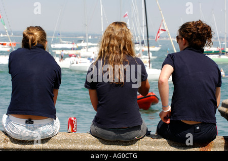 Trois jeunes femmes femmes filles assis sur la digue en regardant le yacht racing au cours de la semaine de Cowes sur l'île de Wight Banque D'Images